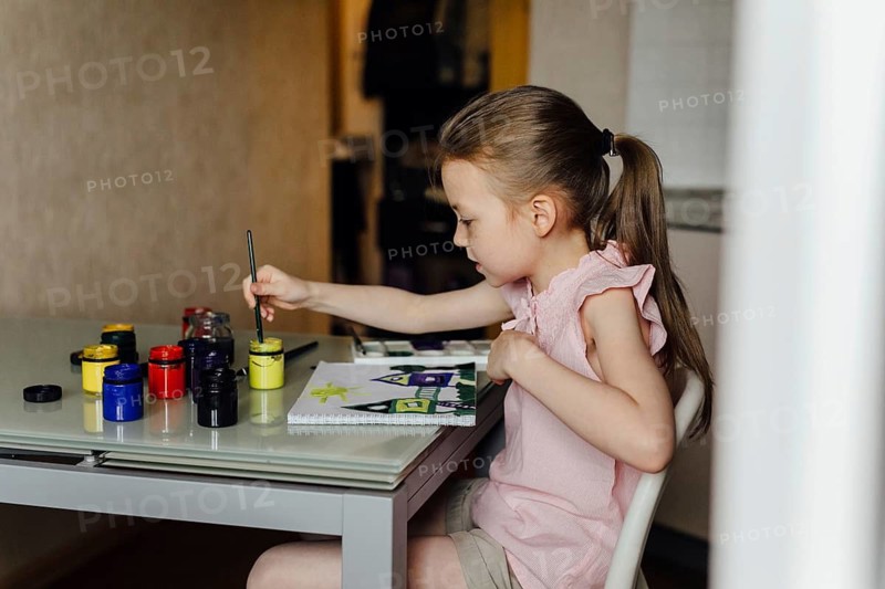 Young girl intently paints with vibrant colors at a home art table, surrounded by paint jars. She exhibits concentration, creativity, and the joy of artistic exploration