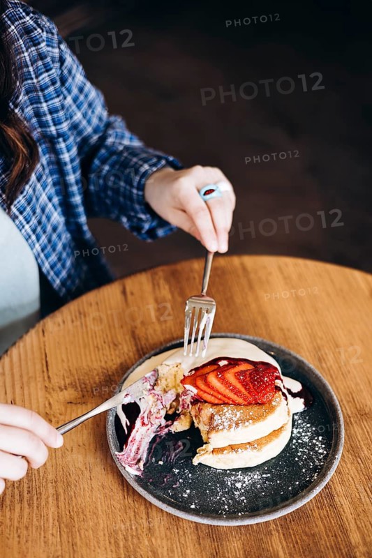 A woman wearing a plaid shirt enjoys fluffy japanese pancakes topped with fresh strawberries and syrup at a cozy cafe. A fork and knife are used on a stylish plate