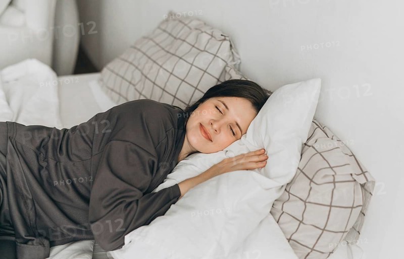 A young woman in comfortable loungewear lies on a white bed, resting her head on soft pillows with a peaceful smile. The cozy bedroom setting creates a warm and relaxing atmosphere