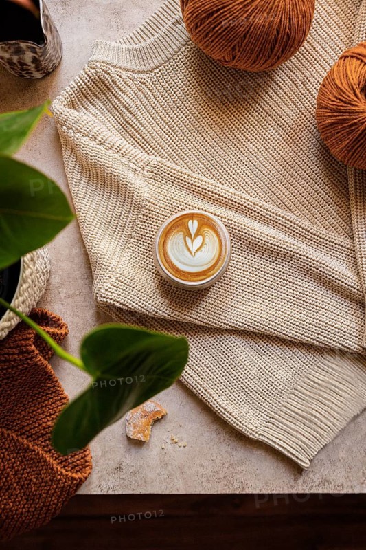 Flat lay of a warm latte on a knitted sweater surrounded by brown yarn balls and green leaves. The cozy setup exudes warmth and tranquility