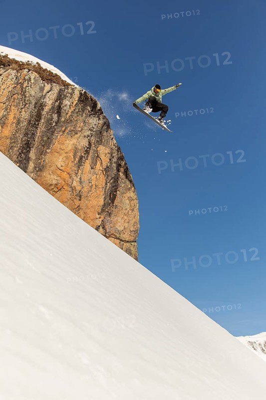 A daring snowboarder executes an exhilarating jump off a snow-covered cliff, soaring high against a clear blue sky in a stunning winter mountain landscape