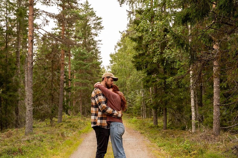 A couple embraces on a scenic path surrounded by lush green trees in a serene Swedish forest. A peaceful moment of connection with nature in a tranquil setting