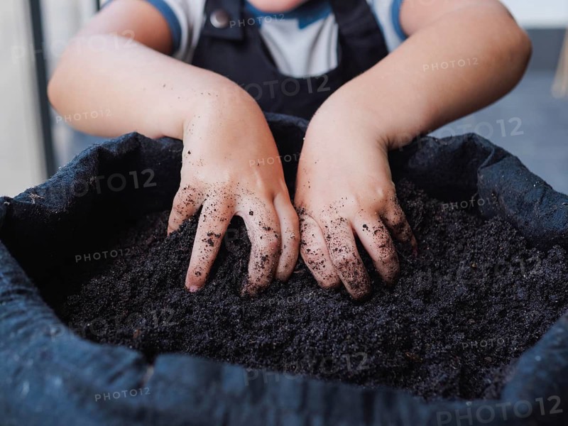 Cropped unrecognizable child with dirt-covered hands is planting seeds in a black grow bag. Kid learning garden work