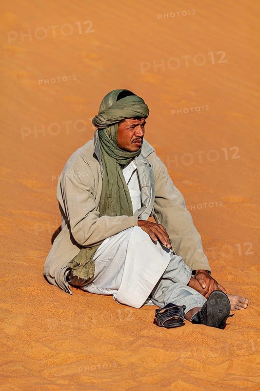 A man sits relaxed on the sand in the desert wearing traditional clothing and a green headscarf, A Tuareg sits in the sand of the Sahara desert in Algeria wearing his traditional garb
