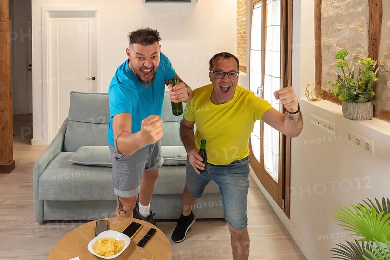 Two male friends celebrating a goal with excitement while watching a sports match at home. Raised fists, beer bottles and strong emotions indoors