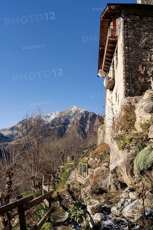 A rustic stone house with a wooden balcony, overlooking a rugged snowy mountain landscape in a serene rural area, under a clear blue sky