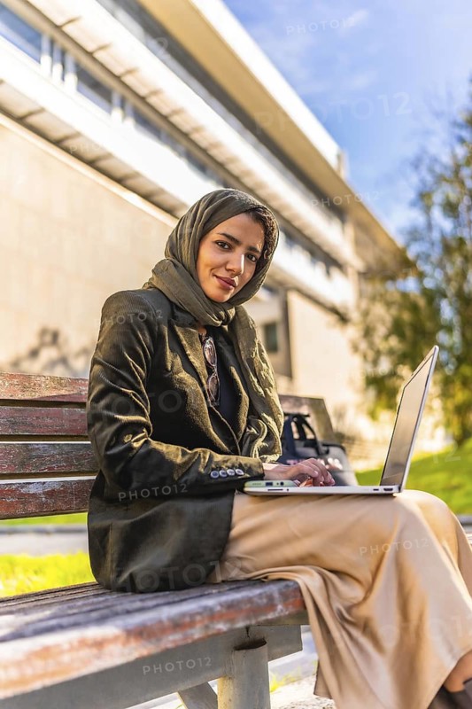 Vertical photo of a young Arab woman in hijab working using laptop sitting on park bench