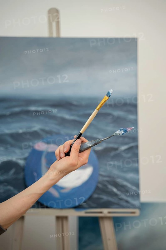 A close up of an artist hand holding a paintbrush and palette knife against a blurred ocean themed canvas. The scene captures the essence of creativity and art in action
