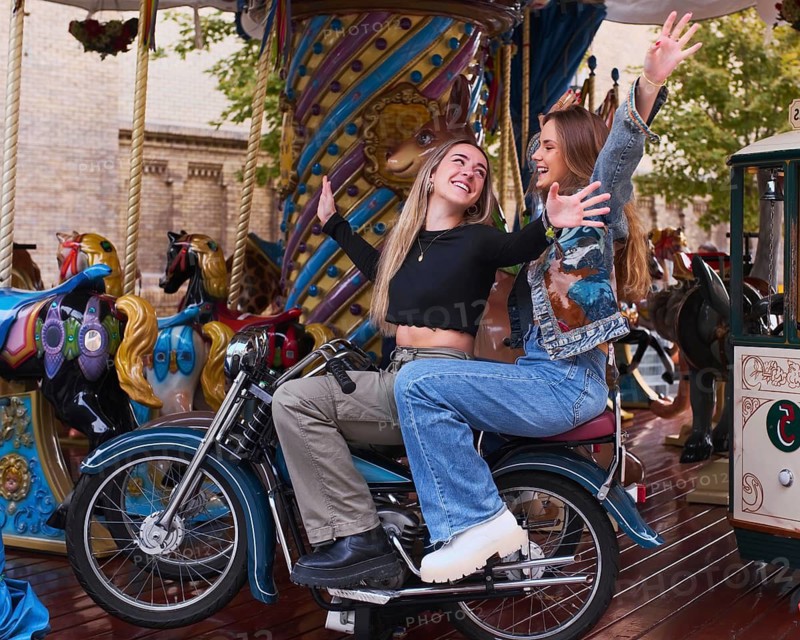 Two smiling friends enjoy a playful moment on a carousel motorcycle in an amusement park, surrounded by colorful horses. Their joyful expressions illuminate the vibrant scene