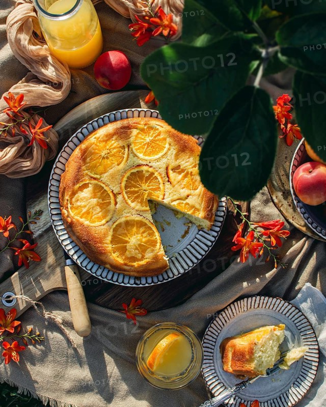 A delicious orange cake topped with fresh orange slices is elegantly displayed with fresh orange juice, apples, and vibrant flowers on a picnic setup in natural light