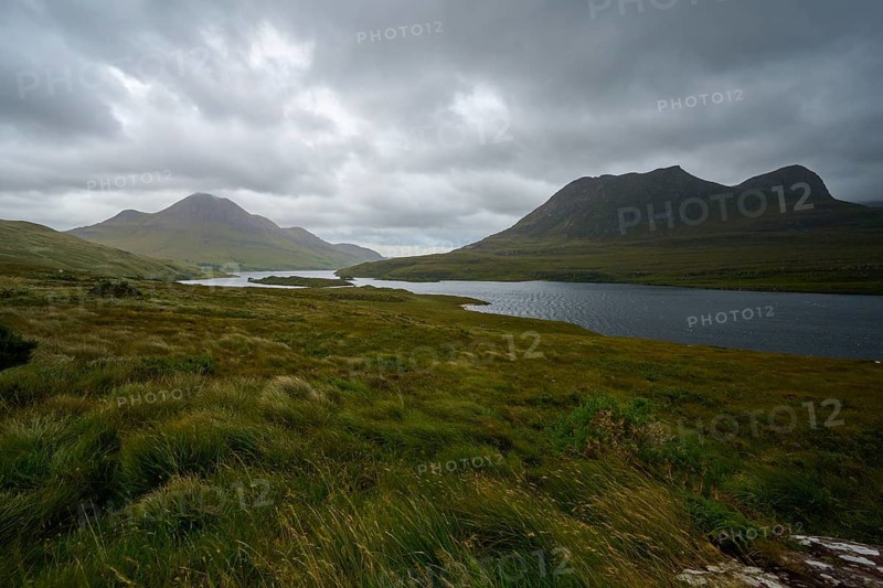 Wide grassland in front of a lake and mountains under a dark, thick sky, Loch Bad a Ghaill, Badnagyle, Ullapool, Scotland, United Kingdom, Great Britain