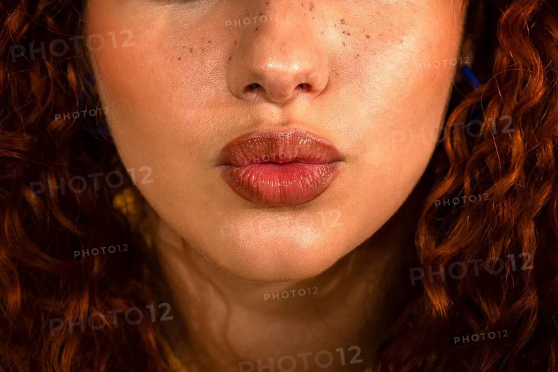 A close-up image captures a woman's glossy lips, slightly pouted, framed by vibrant red curly hair. The freckles around her nose add a touch of natural beauty