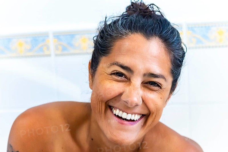 A joyful woman with a radiant smile, captured in natural light on a bathroom. Her expressive eyes and laughter convey happiness and warmth, set against a soft, neutral background at home