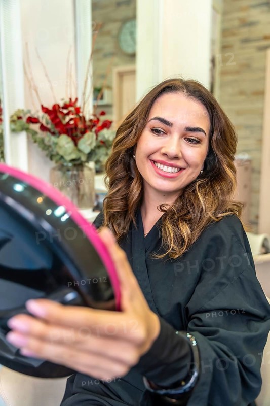 Young woman smiling, looking at her reflection in a handheld mirror, showing satisfaction with her wavy, brown hair after a beauty care makeover at a modern salon
