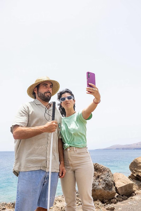 A blind man with a cane and a woman stand close together, capturing a selfie by the coast. The sunny ocean backdrop highlights their shared moment of adventure and connection