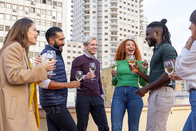 A lively group of friends celebrate on a rooftop, holding glasses of white wine They share laughter and conversation, surrounded by a cityscape with tall buildings