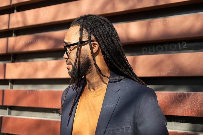 A cuban business man with long dreadlocks, wearing sunglasses and a blazer, basks in the sunlight. The ambient light and wood backdrop create a warm atmosphere