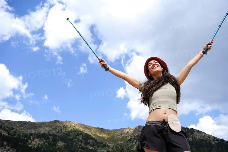 A joyful woman with an ostomy bag raises hiking poles, embracing nature and freedom in a scenic outdoor setting Celebrating empowerment and resilience in the great outdoors