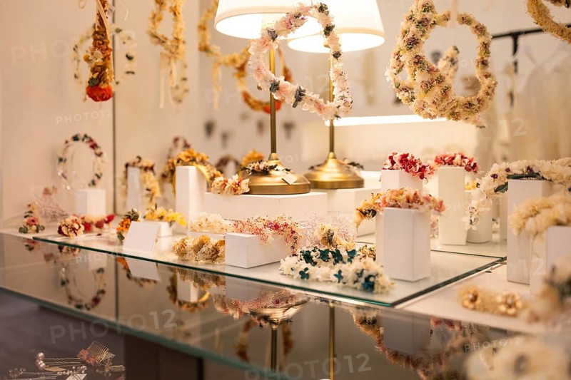 A variety of handcrafted floral headpieces are elegantly displayed on glass shelves. Soft lighting accentuates the intricate details and vibrant colors, creating a warm ambiance