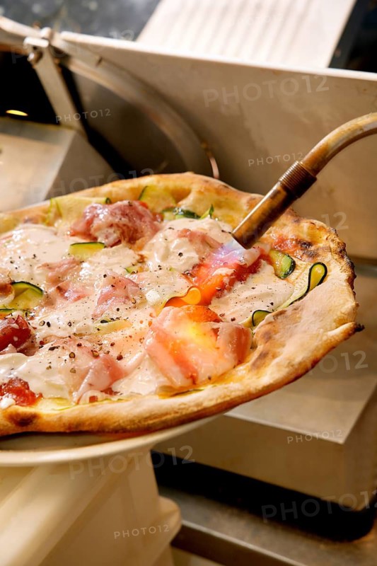 A freshly baked pizza topped with zucchini, bacon, and a melted cheese blend is served hot straight from the oven using a wooden pizza peel