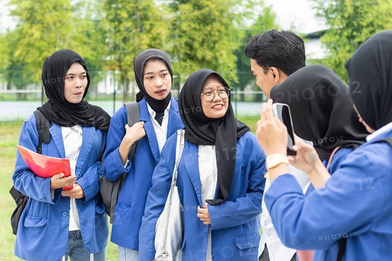 A group of Asian Muslim students, wearing hijabs and matching blue uniforms, gather outdoors with backpacks and papers, happily chatting and interacting before class
