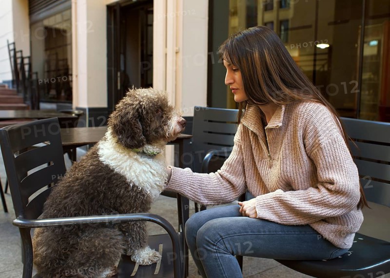 A young woman lovingly interacts with her dog while sitting in an outdoor urban cafe setting. Both are engaged in a moment of connection, captured in a candid photo