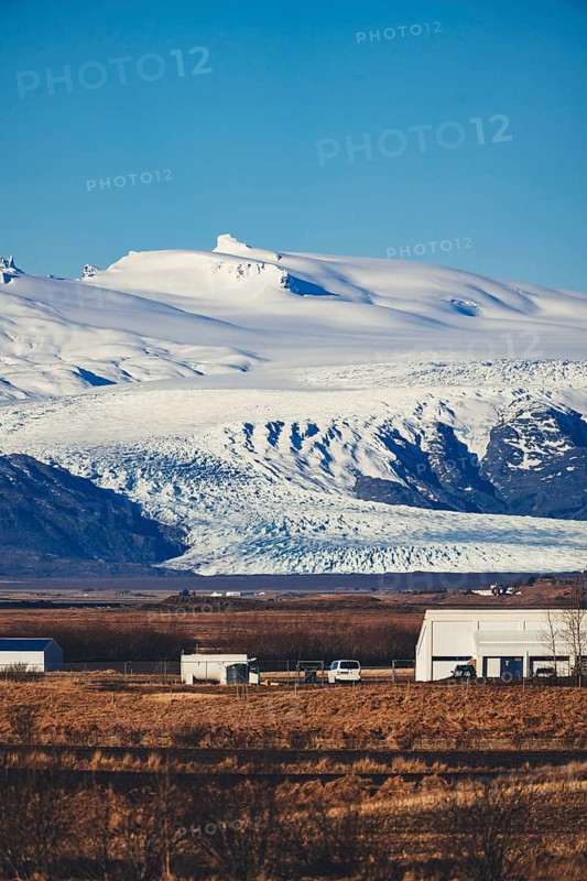 Distant view of a glacier from the Stokksnes area in Iceland, with small rural buildings and golden fields in the foreground under a bright blue sky