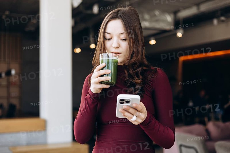 A young female student in a casual setting takes a break while sipping a green smoothie and browsing her smartphone, perfectly captured in a moment of modern-day multitasking