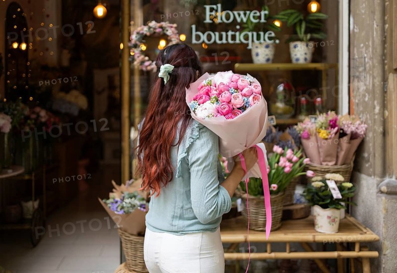 A woman stands in a flower shop, holding a vibrant bouquet of pink and white flowers. The shop is beautifully decorated with various floral arrangements, creating a warm and inviting atmosphere