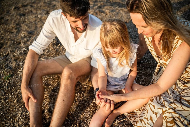 A loving family sits together on a sandy beach in Italy, sharing a warm and peaceful moment. The parents gently hold their daughter's hands as they enjoy their time outdoors