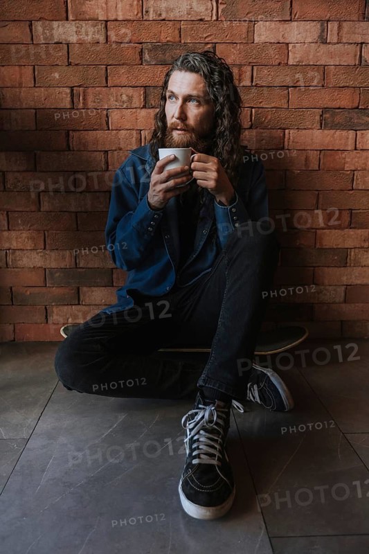 A man with long hair and a beard sits on the floor against a brick wall, holding a cup of coffee. He appears deep in thought, wearing casual clothing and sneakers