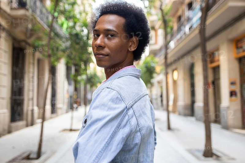 A non-binary person of African American descent walks confidently down a sunlit urban street, wearing a light denim jacket. The scene captures diversity and individuality