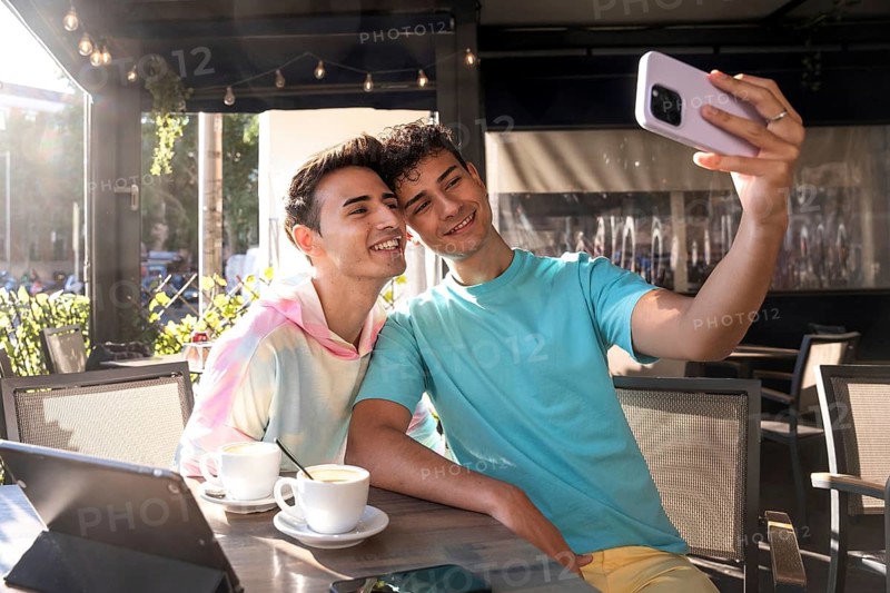 A cheerful gay couple enjoys sunny moments, taking a selfie together at an outdoor cafe. Relaxed atmosphere with coffee cups and digital tablet on the table