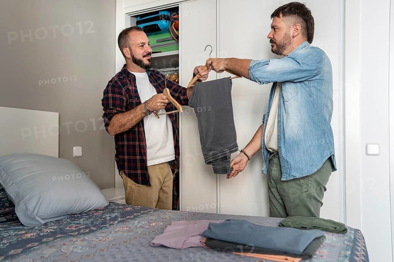 A gay couple stands in their bedroom, selecting clothes from a wardrobe. One holds a pair of pants on a hanger while the other assists, surrounded by neatly folded clothing on the bed