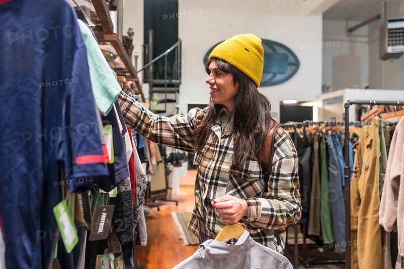 Young woman smiling while browsing clothing racks and holding a hanger in a thrift boutique, exploring sustainable, secondhand fashion choices and casual urban style finds