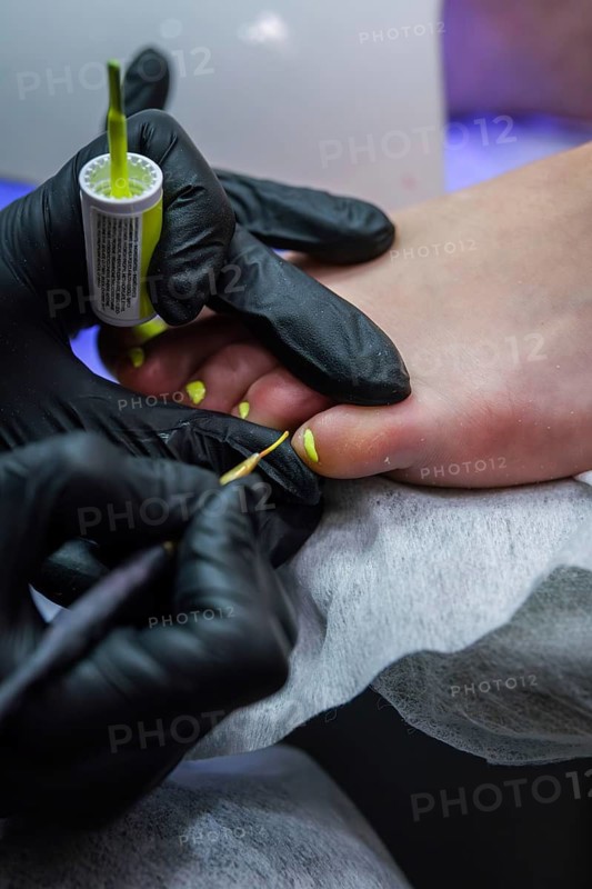 Vertical closeup of toenail polish being applied during a professional pedicure, with gloved hands shaping and detailing the nails on a resting foot