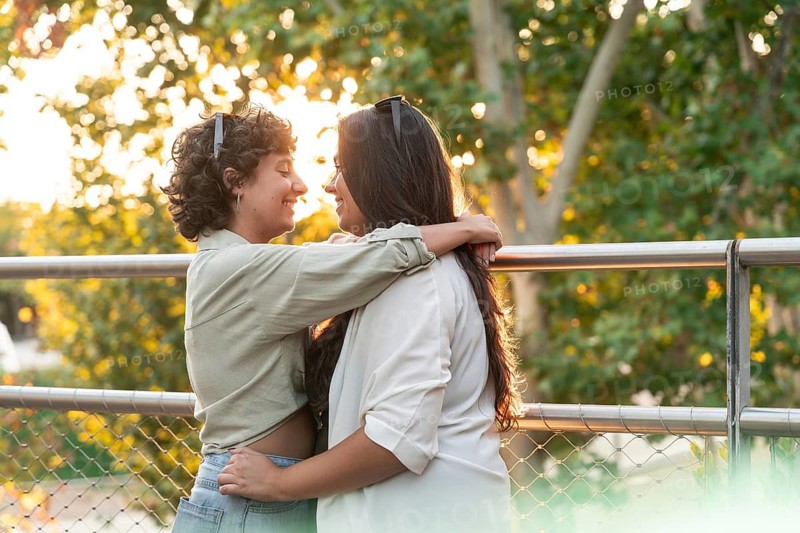 A lesbian couple shares a romantic embrace in a sunlit park. They look into each other's eyes, surrounded by vibrant greenery, capturing a tender moment of love and connection