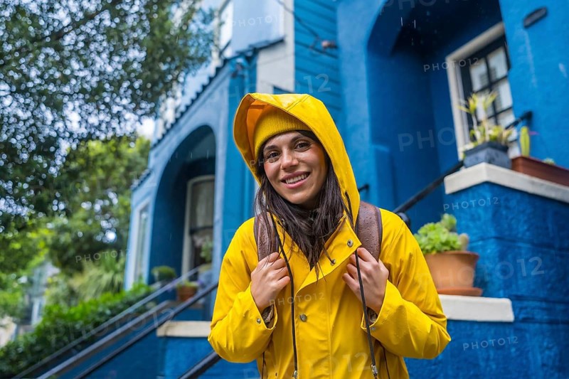 Woman standing outdoors during rainy weather, wearing a yellow beanie and rain jacket, holding hands to her collar, with a vibrant blue building in the background