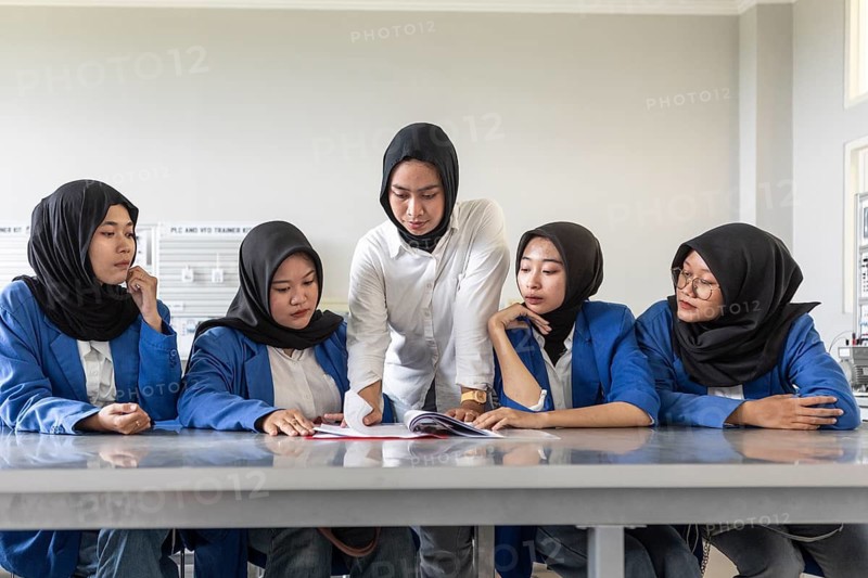 Asian Muslim teacher instructing a group of attentive students in a classroom setting. The students, wearing headscarves, focus intently on a textbook as the teacher explains