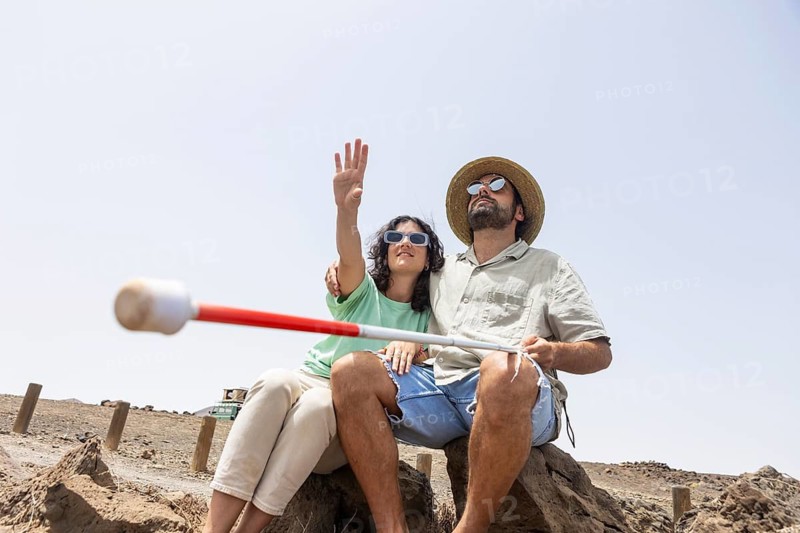 A blind man and a woman sit on rocks in the desert, with the woman pointing towards the horizon. The moment highlights teamwork, exploration, and their adventurous spirit