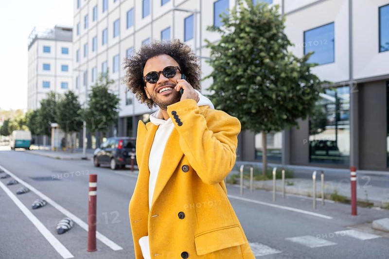 A businessman with afro hair wearing a stylish yellow coat and sunglasses, happily talking on the phone while walking through a modern urban street