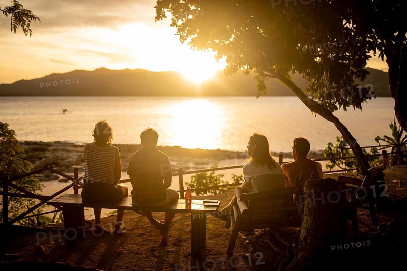 Back view of unrecognizable friends sitting together on a bench, enjoying a breathtaking sunset over the sea in the Philippines