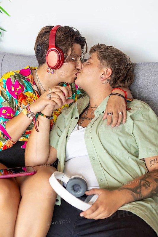 A lesbian couple sitting on a sofa, sharing a kiss while holding hands. One wears vibrant red headphones, indicating they are enjoying music together in home routines