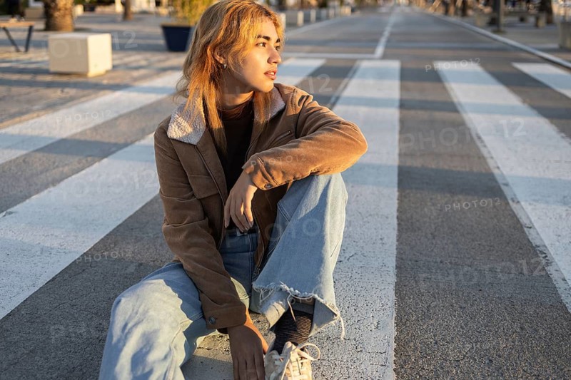 A Mixed-race woman wearing a brown jacket and jeans sits casually on a crosswalk, gazing into the distance The setting is outdoor and urban, captured during sunset