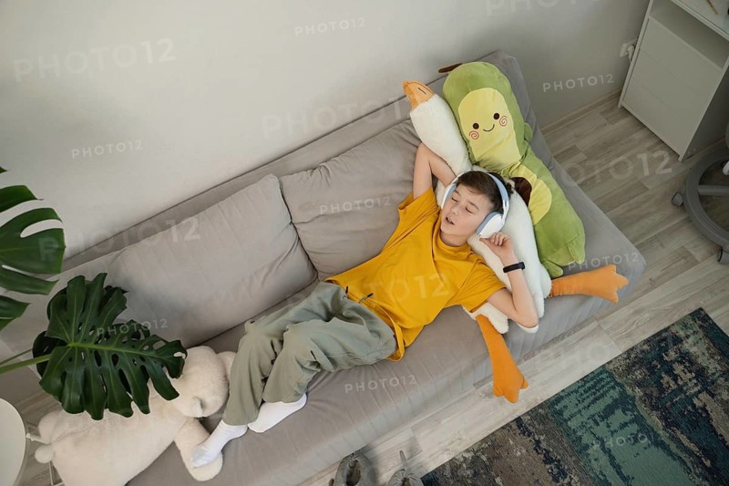 A young boy in a yellow shirt, wearing headphones, relaxes on a sofa with stuffed toys. The cozy setting includes plush cushions and decorative plants