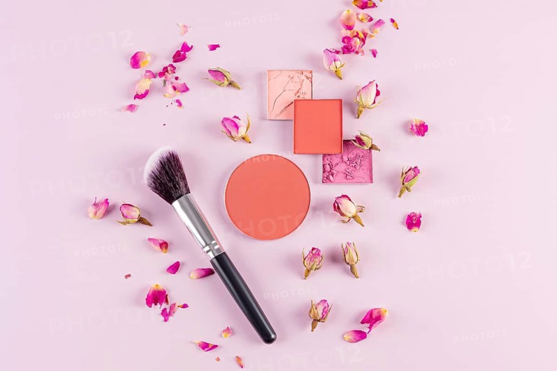 A flat lay composition of makeup essentials including blushes, a makeup brush, and scattered rose petals on a soft pink background, showcasing beauty and elegance