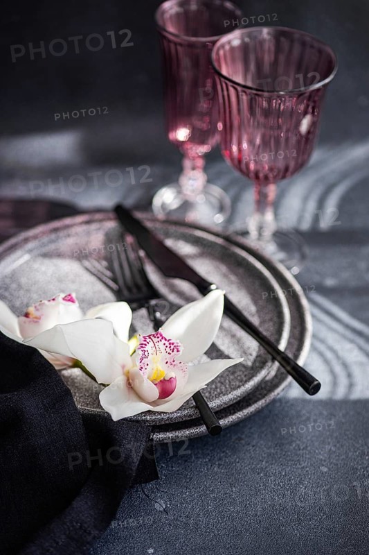 A sophisticated table setting featuring textured plates, dark cutlery, and delicate white orchids. Two pink glasses enhance the romantic ambiance against a soft background