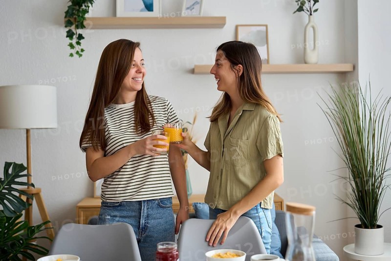A lesbian couple shares a joyful moment at home, clinking glasses of orange juice Surrounded by cozy decor, they celebrate love and togetherness LGBTQIA+ pride