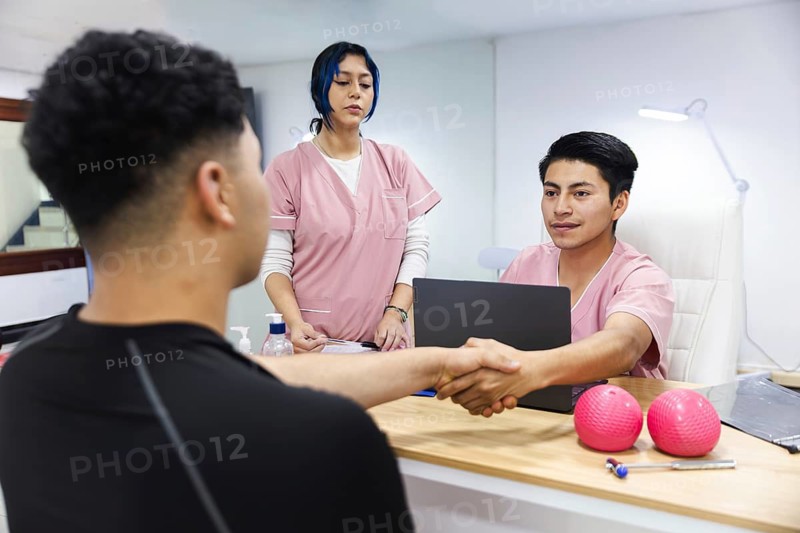 In an Ecuadorian physiotherapy clinic, a patient shakes hands with a smiling therapist. An attentive staff member observes, creating a welcoming ambiance. Treatment tools nearby
