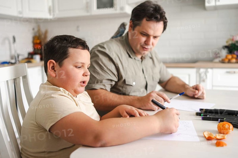A father and son sit together at a kitchen table drawing, while a bird perches nearby. The atmosphere is warm and cozy, highlighting family bonding and creativity